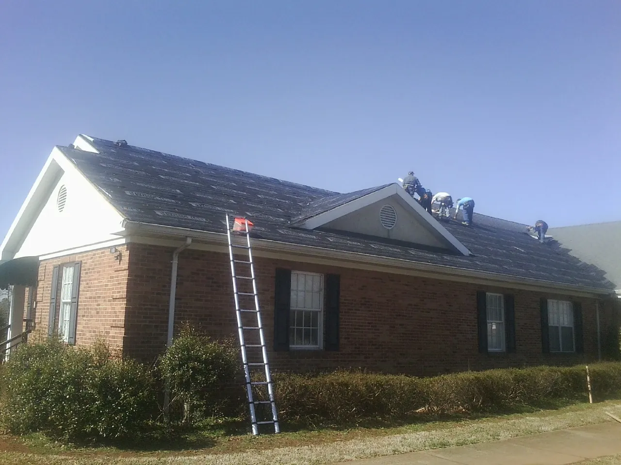 Crew tearing shingles off a brick ranch roof with a ladder set — Emergency Roof Repair work by our crew in Westhampton
