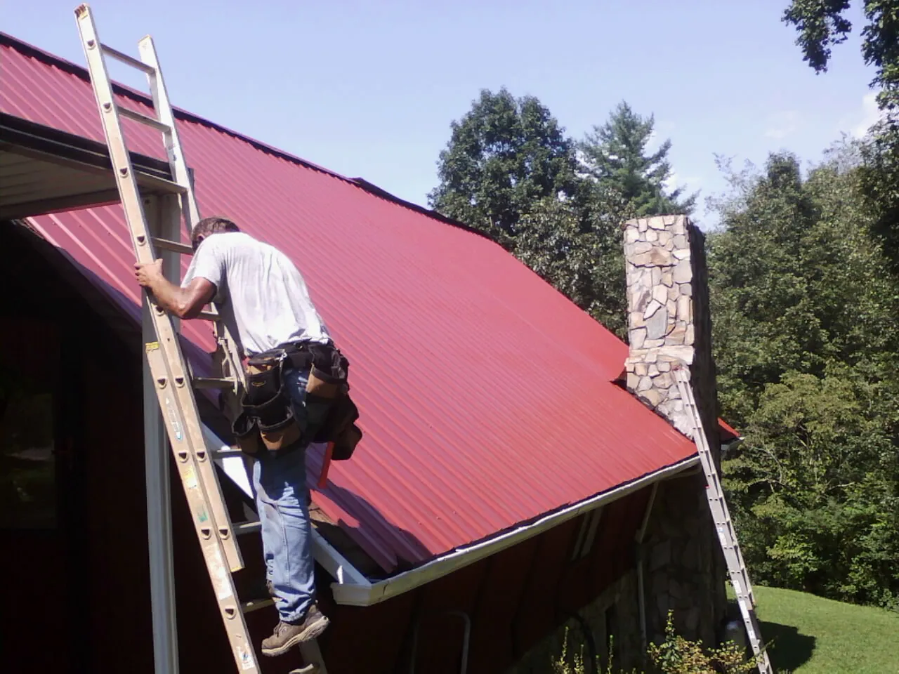 Roofer on a ladder beside a red metal roof and stone chimney — Soffit & Fascia Repair by Roofing Brothers in Westhampton